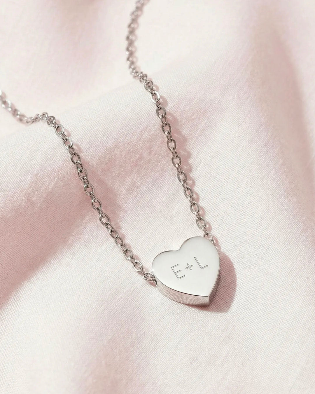 Silver heart-shaped necklace with 'E+L' initials on a pink background